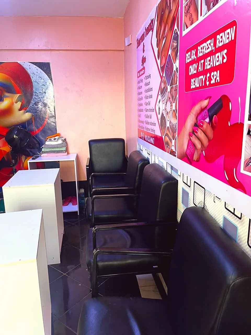 Row of black client chairs beneath the salon's pink services poster, with nail tables and a hood dryer